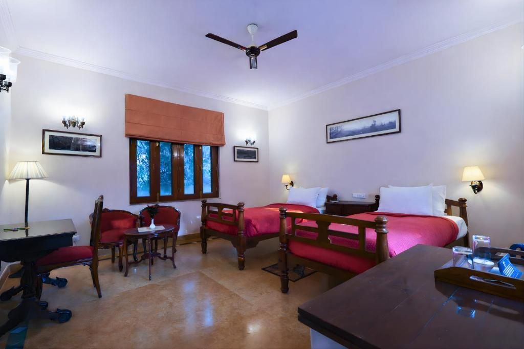 A hotel room with two beds, a desk, a chair, a table, a ceiling fan, and a window with blinds.