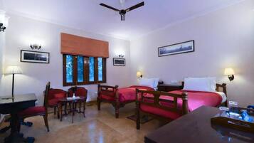 A hotel room with two beds, a desk, a chair, a table, a ceiling fan, and a window with blinds.