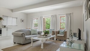 Una sala de estar con un sofá, una mesa de centro y una silla de mimbre. Hay ventanas con cortinas, una chimenea y un televisor sobre un mueble.