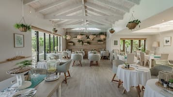 Un comedor espacioso con techos altos y plantas colgantes, con mesas dispuestas para comer y sillas alrededor.