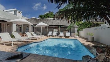 A swimming pool with lounge chairs and umbrellas, surrounded by a thatched-roof building and lush greenery.