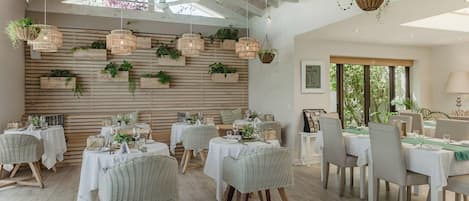 A dining area with multiple tables, wicker chairs, hanging planters, and a wooden ceiling structure.