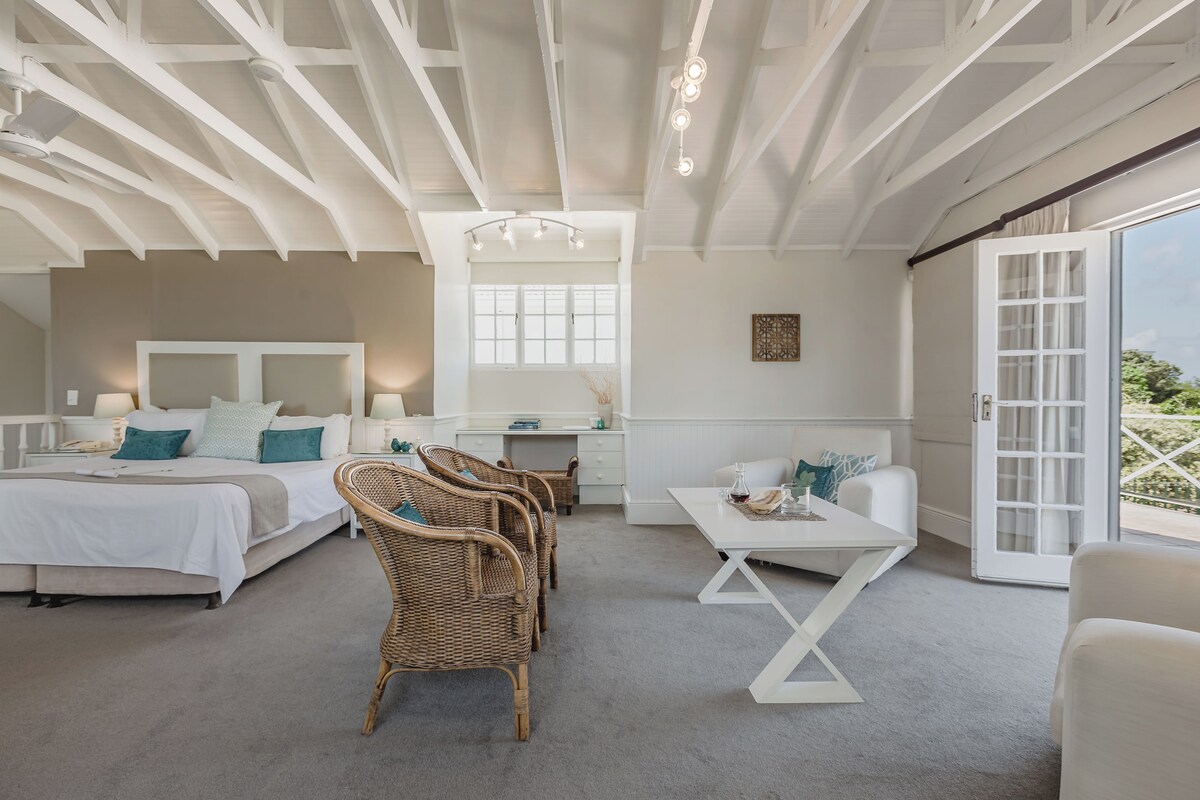 A spacious bedroom with a large bed, a desk, wicker chairs, and a balcony with a view.