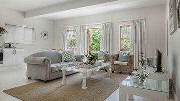 A modern living room with a sofa, coffee table, and wicker chairs. There is a kitchen area with cabinets and a window.