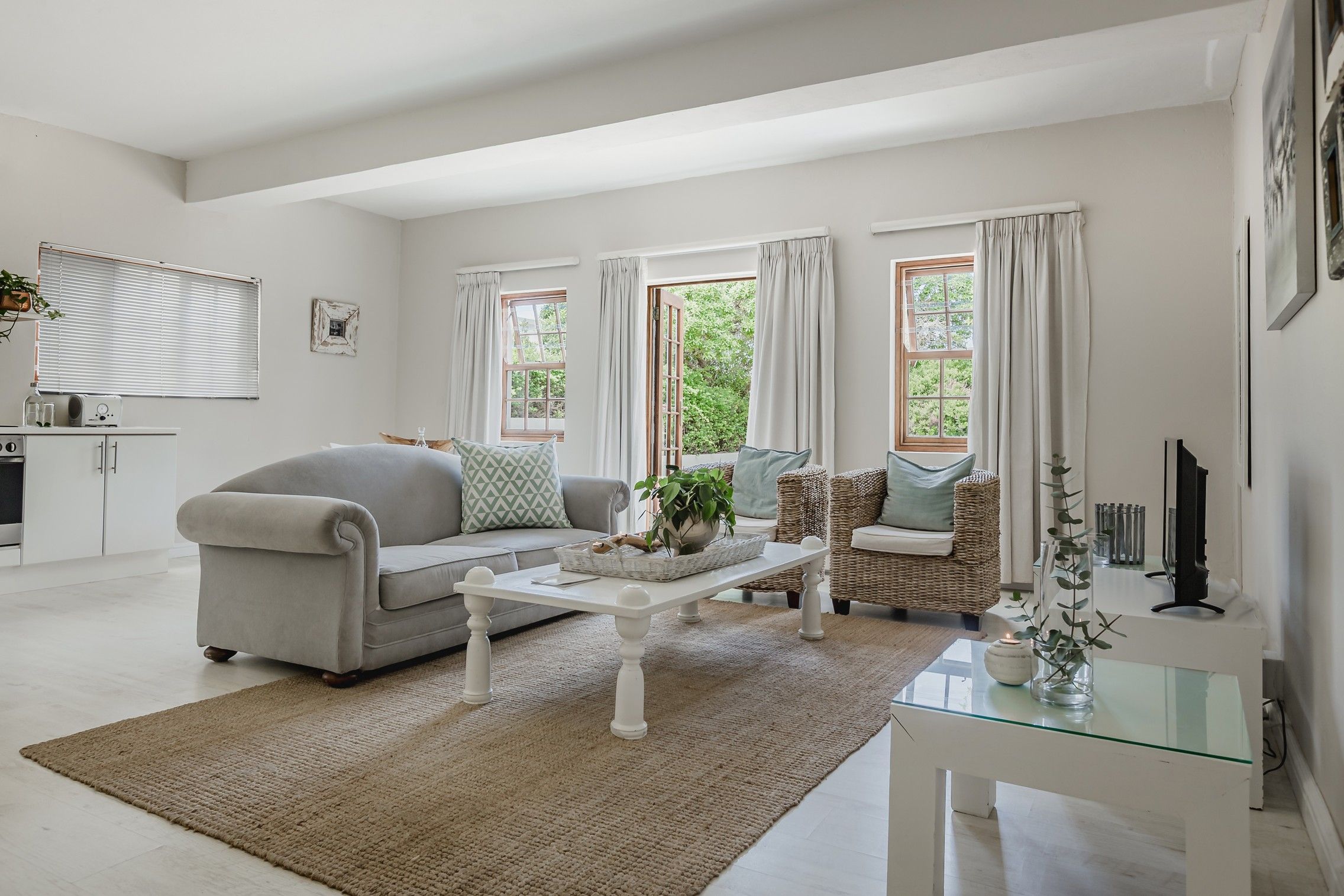Una sala de estar con un sofá, una mesa de centro y una silla de mimbre. Hay ventanas con cortinas, una chimenea y un televisor sobre un mueble.