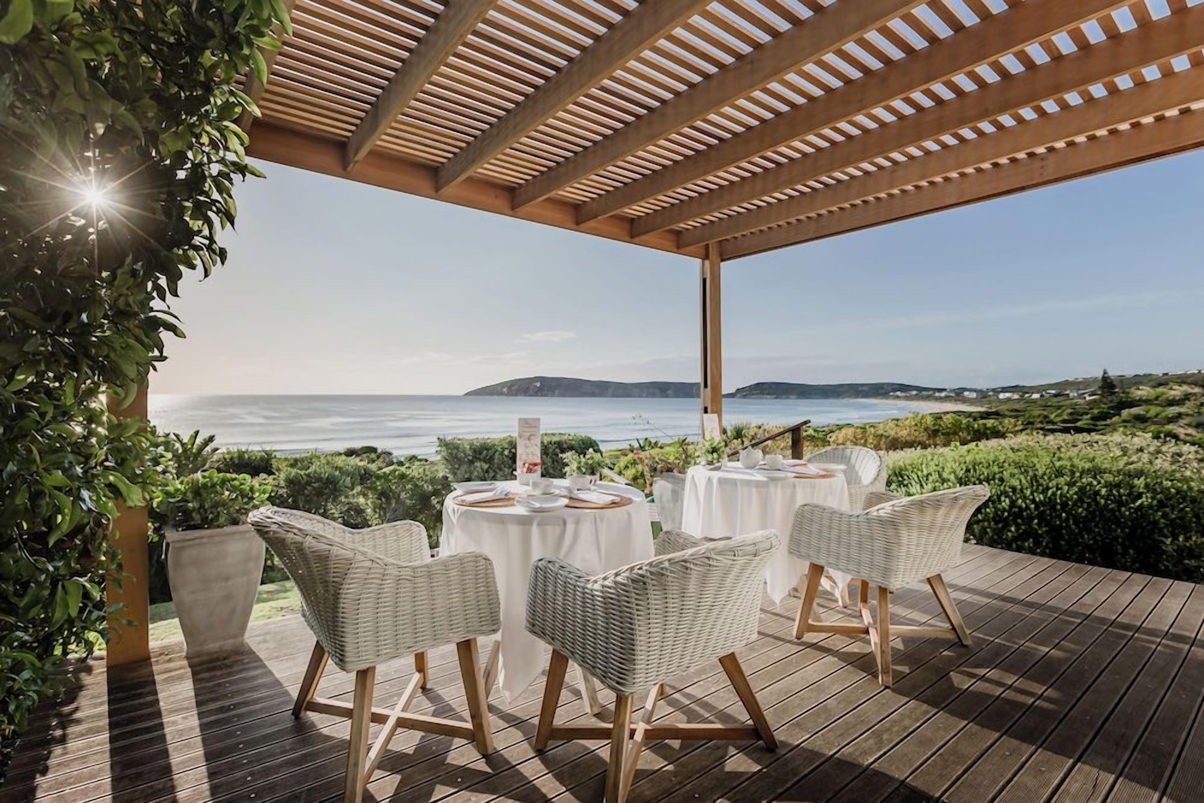 Un área de comedor al aire libre con sillas de mimbre y mesas redondas, con vistas a un paisaje costero.