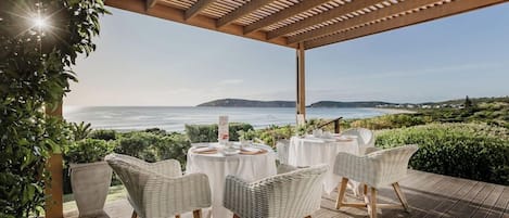 Un área de comedor al aire libre con sillas de mimbre y mesas redondas, con vistas a un paisaje costero.