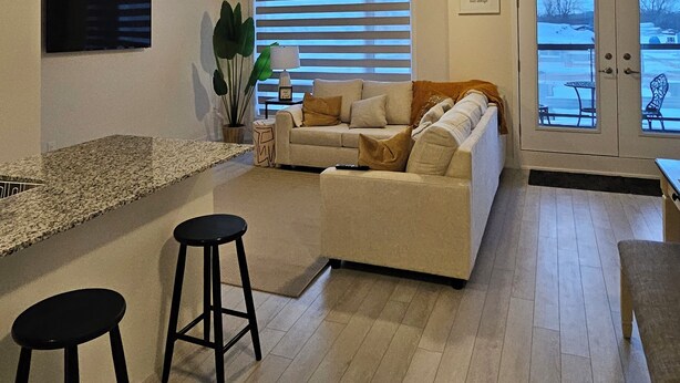 Una sala de estar moderna con un sofá, dos taburetes altos, un televisor y una ventana con persianas.