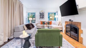 Ein modernes Wohnzimmer mit Sofa, Sessel und Couchtisch, einem Kamin und einem an der Wand montierten Fernseher.
