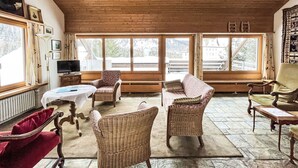 Ein gemütliches Zimmer mit Holzboden, großen Fenstern, Korbmöbeln und einem Fernseher.