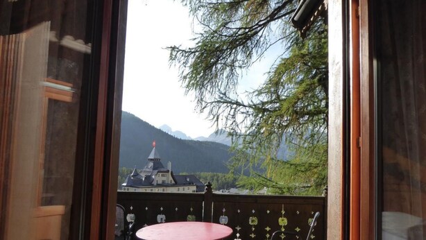 A room with a view of a mountain and a building. There is a table and chairs in the foreground.