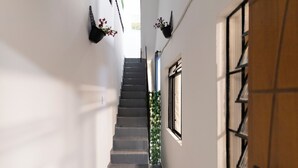 Ένα στενό κλιμακοστάσιο με μια ξύλινη πόρτα στα δεξιά, ένα παράθυρο με μαύρα μεταλλικά κάγκελα και μια κρεμαστή γλάστρα στον τοίχο με λουλούδια.