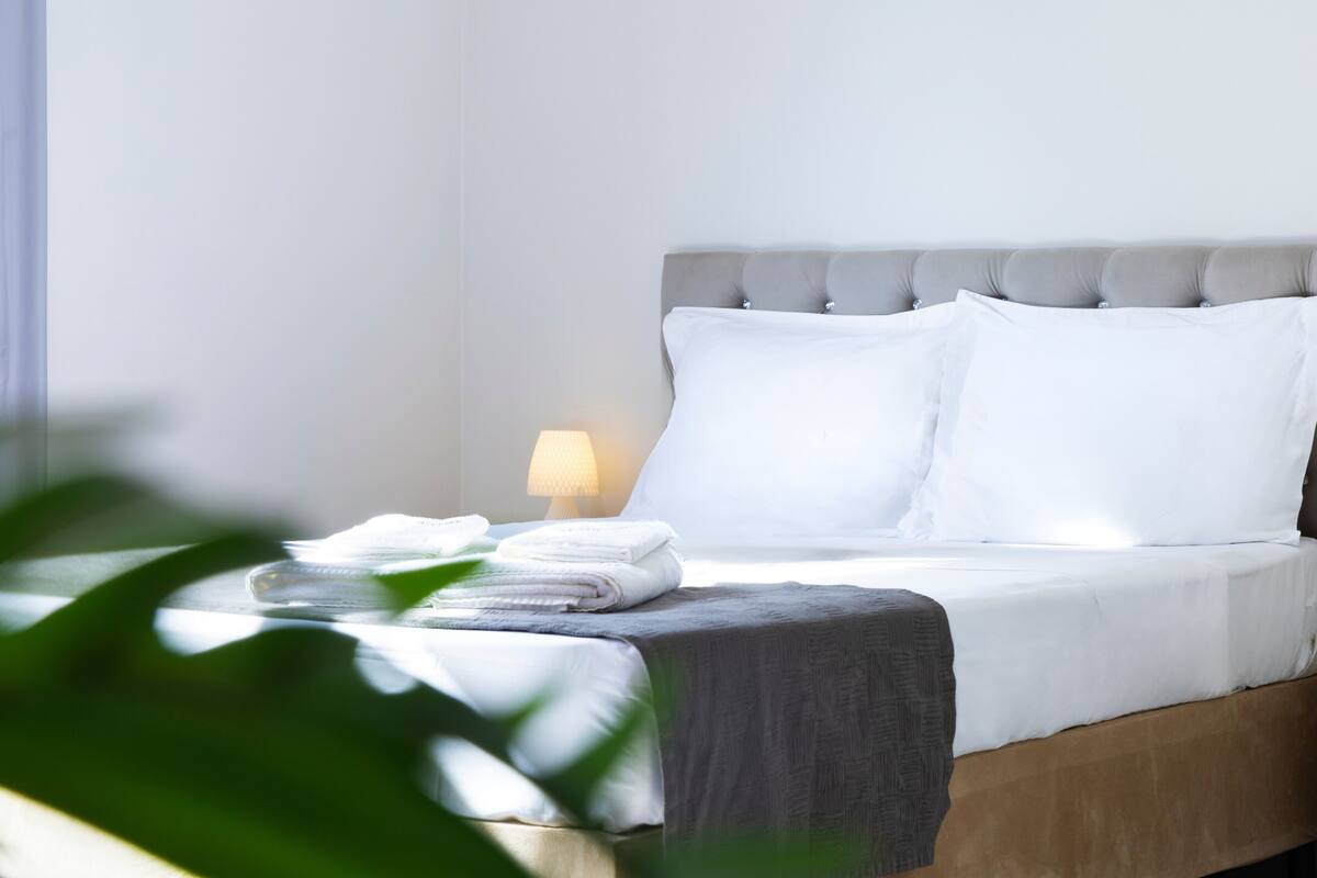 A neatly made bed with white linens, a grey headboard, and a bedside lamp.