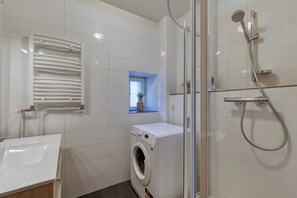 Une salle de bain moderne dotée d’un lave-linge encastré, d’une cabine de douche en verre et d’un mur recouvert de carreaux blancs.
