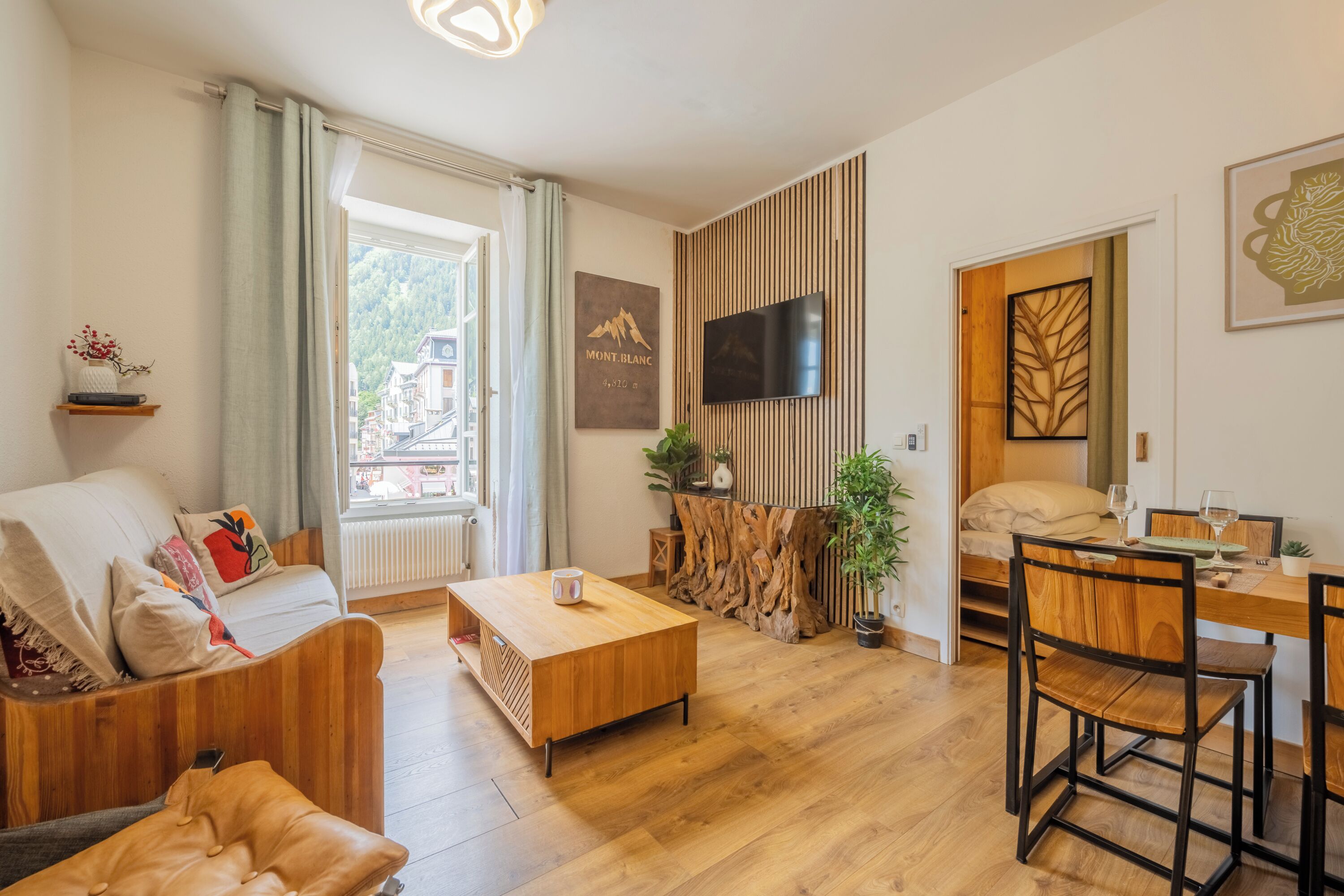 Un salon moderne comprenant un canapé, une table basse, une table à manger et des chaises. Un téléviseur est fixé au mur, on y trouve également un morceau de tronc d’arbre en bois et une fenêtre donnant sur des immeubles.