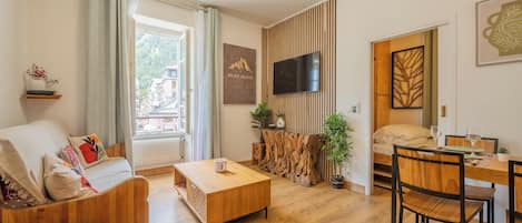 Un salon moderne comprenant un canapé, une table basse, une table à manger et des chaises. Un téléviseur est fixé au mur, on y trouve également un morceau de tronc d’arbre en bois et une fenêtre donnant sur des immeubles.
