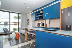 Une cuisine moderne avec des meubles de couleur bleue, un évier en acier inoxydable et une machine à café. On y trouve également un coin bar avec des tabourets et une vue sur l’extérieur grâce à de grandes fenêtres.
