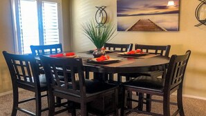 Un coin repas avec une table ronde, des chaises noires, un vase avec des plantes vertes et un tableau représentant un coucher de soleil accroché au mur.