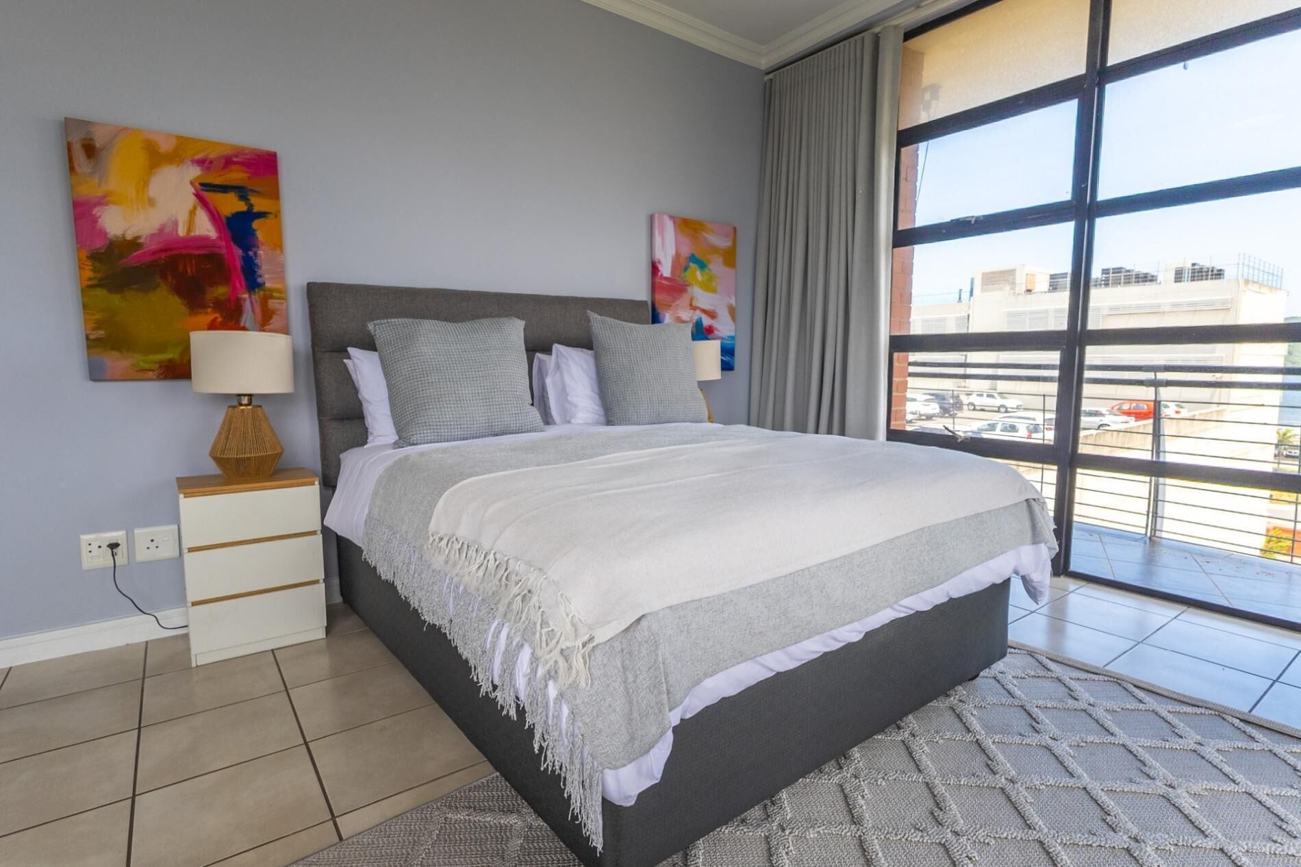 Une chambre à coucher moderne avec un grand lit, une table de chevet, une lampe et deux tableaux abstraits accrochés au mur.