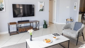 Ein modernes Wohnzimmer mit Flachbildfernseher, Couchtisch mit Vase und Sushi, grauem Sessel und einer Holzbank.