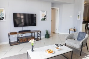 Ein modernes Wohnzimmer mit Flachbildfernseher, Couchtisch mit Vase und Sushi, grauem Sessel und einer Holzbank.