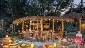 Un área de bar al aire libre con mobiliario de madera, techo de paja y luces colgantes.
