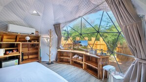 Habitación tipo tienda con cama, estantes de madera, escritorio y vistas a través de un ventanal grande.