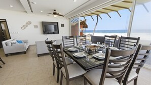 Un coin repas avec une grande table dressée pour six personnes, entourée de chaises, d’un téléviseur et offrant une vue sur la plage.