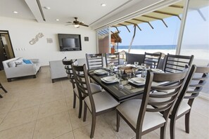 Un comedor con una mesa grande dispuesta para seis personas, rodeada de sillas, un televisor y vistas a la playa.
