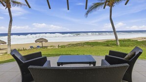 Un espace extérieur aménagé avec des meubles en osier et une table, offrant une vue sur une plage avec des vagues et un ciel dégagé.
