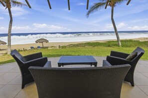 An outdoor seating area with wicker furniture and a table, overlooking a beach with waves and a clear sky.