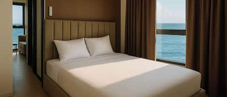 A large bed with a headboard, a view of the sea through a window, and a door leading to another room.