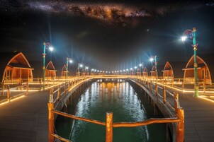 Vista nocturna de un paseo marítimo con pabellones de madera y una superficie acuática reflectante.