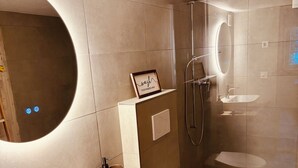 A modern bathroom with a large oval mirror, a rectangular sink, a toilet, and a glass-enclosed shower area. - Beautiful, gently renovated apartment in the beautiful Bernese Oberland. (Frutigen)