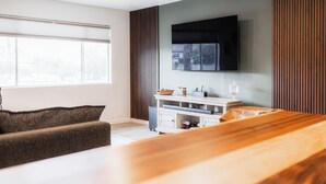 Ein modernes Wohnzimmer mit einem Holzesstisch, einer Couch, einem an der Wand montierten Fernseher und einem Fenster mit Jalousien.