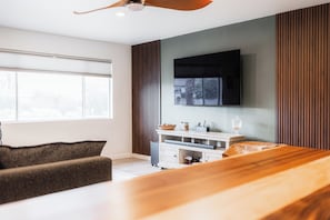 Una sala de estar moderna con una mesa de comedor de madera, un sofá, un televisor montado en la pared y una ventana con persianas.