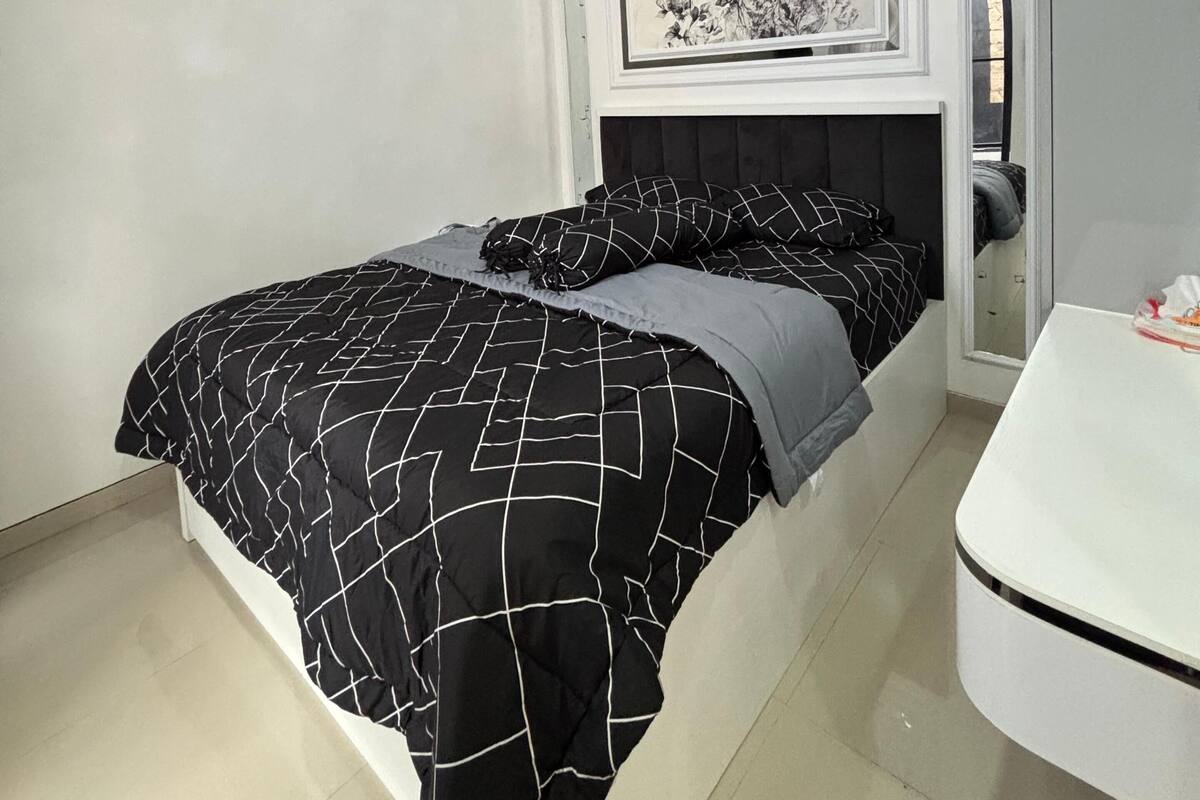 A bedroom with a bed, a white desk, an air conditioning unit, and a ceiling light fixture.