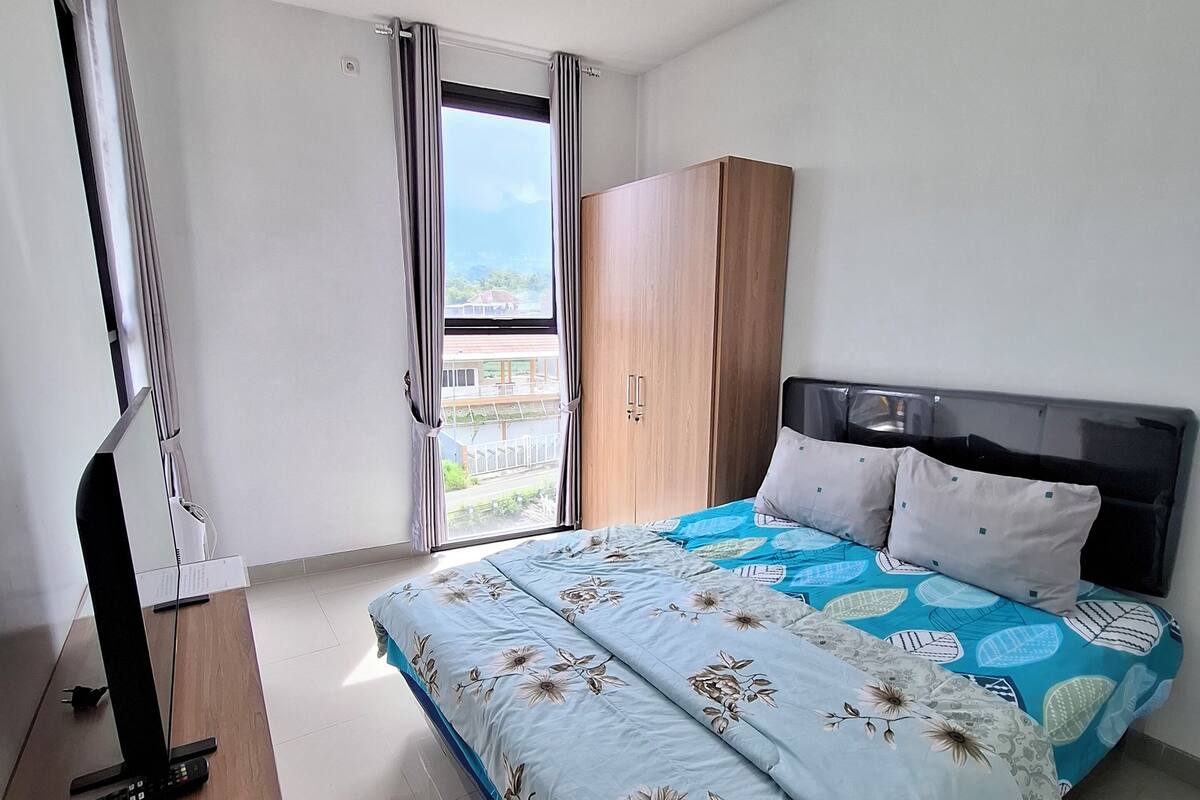 A bedroom with a bed, a wardrobe, a desk with a TV, and a window with curtains.