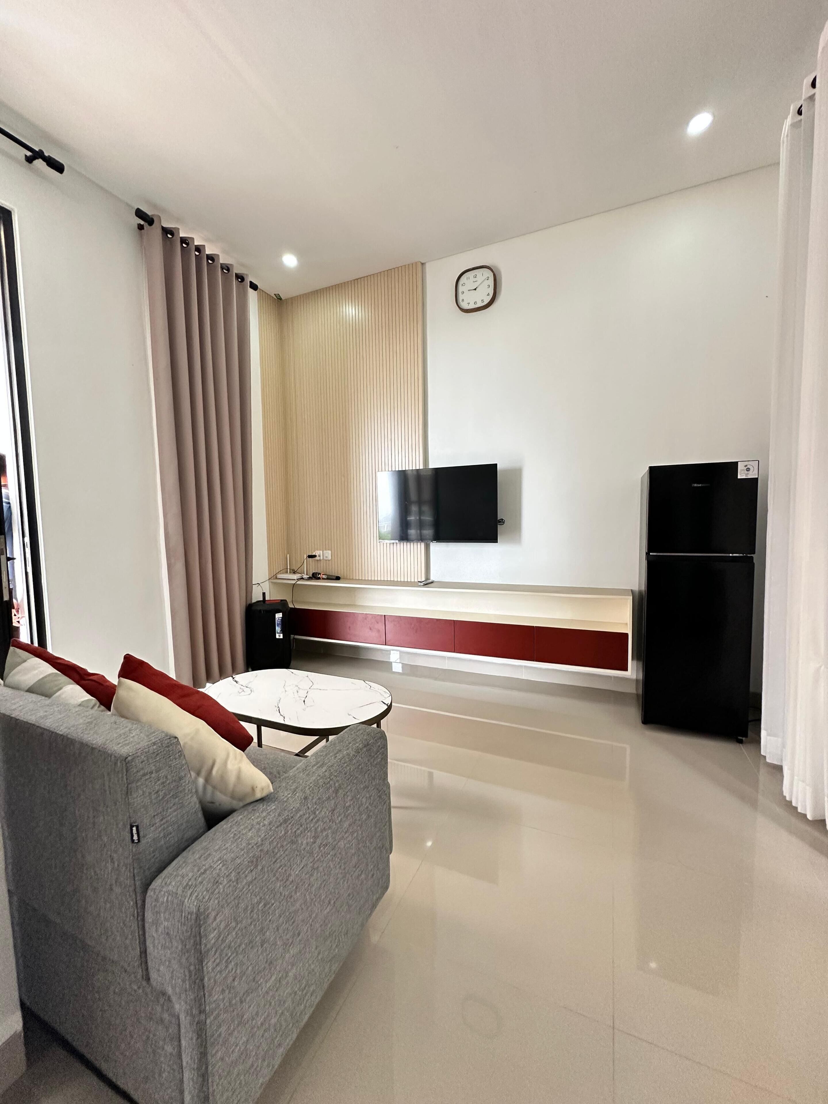 A modern living room with a sofa, a flat-screen TV mounted on the wall, a small table, and a refrigerator.