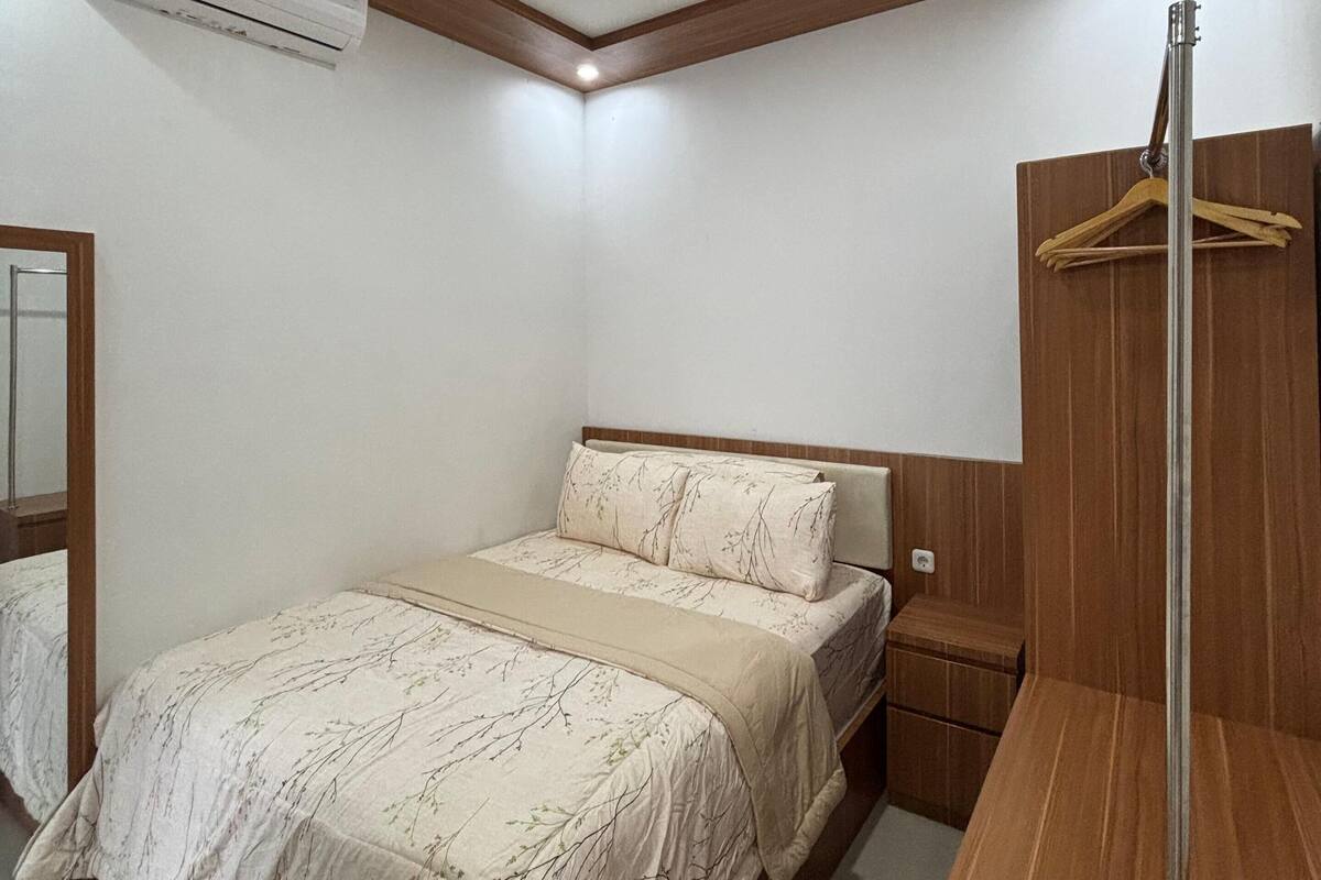 A bedroom with a wooden headboard, a bed with a beige floral bedspread, an air conditioning unit, a wooden nightstand, and a wooden wardrobe with a hanger.