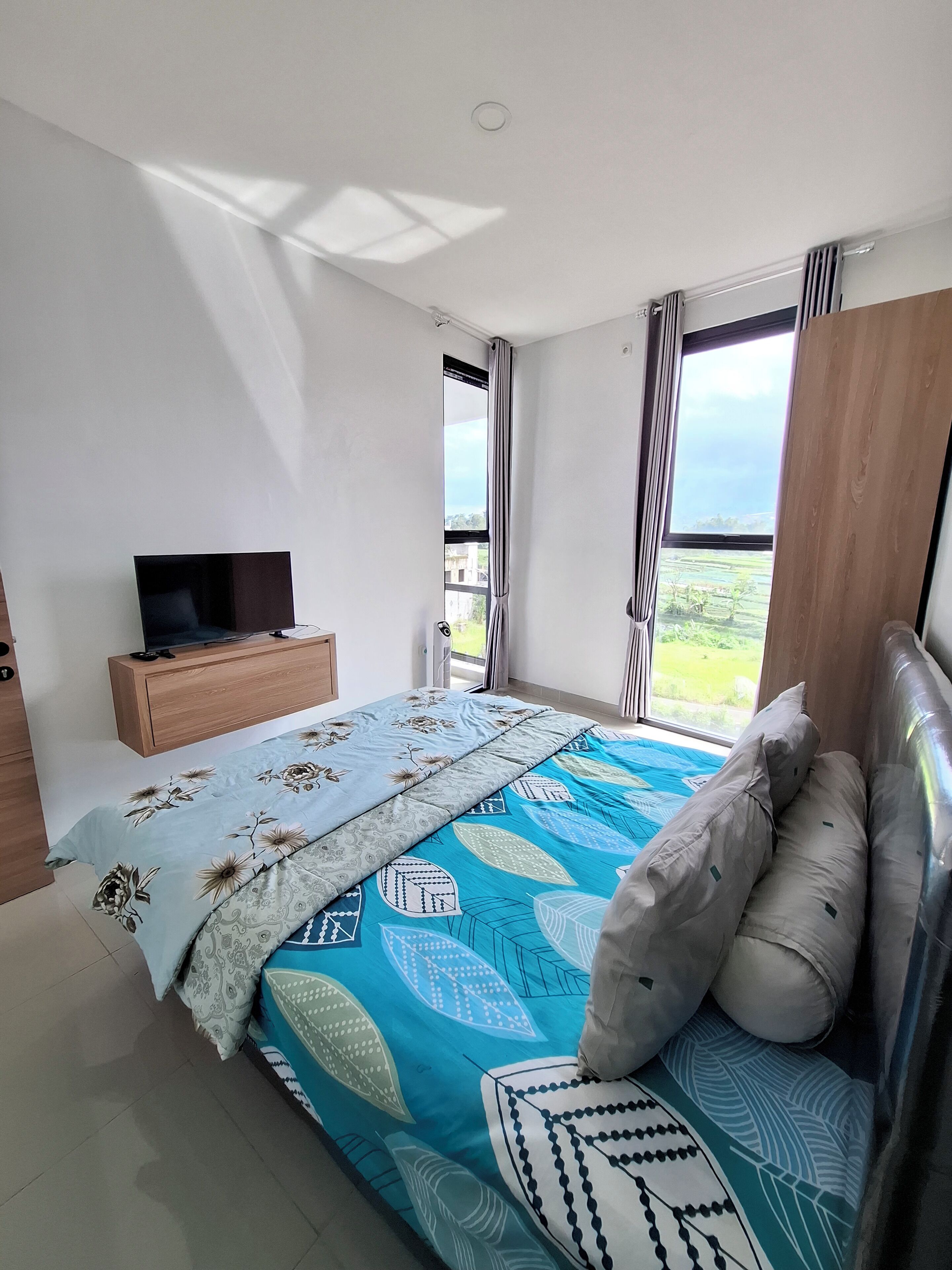 Un dormitorio con una cama, un televisor sobre un mueble de madera y un ventanal con cortinas.