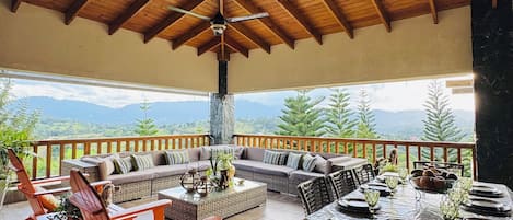 A covered outdoor seating area with wooden furniture, a dining table set for six, and a view of mountains in the background.