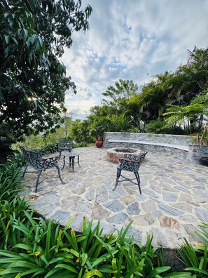 An outdoor patio with stone flooring and metal chairs, surrounded by lush greenery and a stone wall. - Luxury Mountain Villa w Pool & Fire Pits (Jarabacoa)