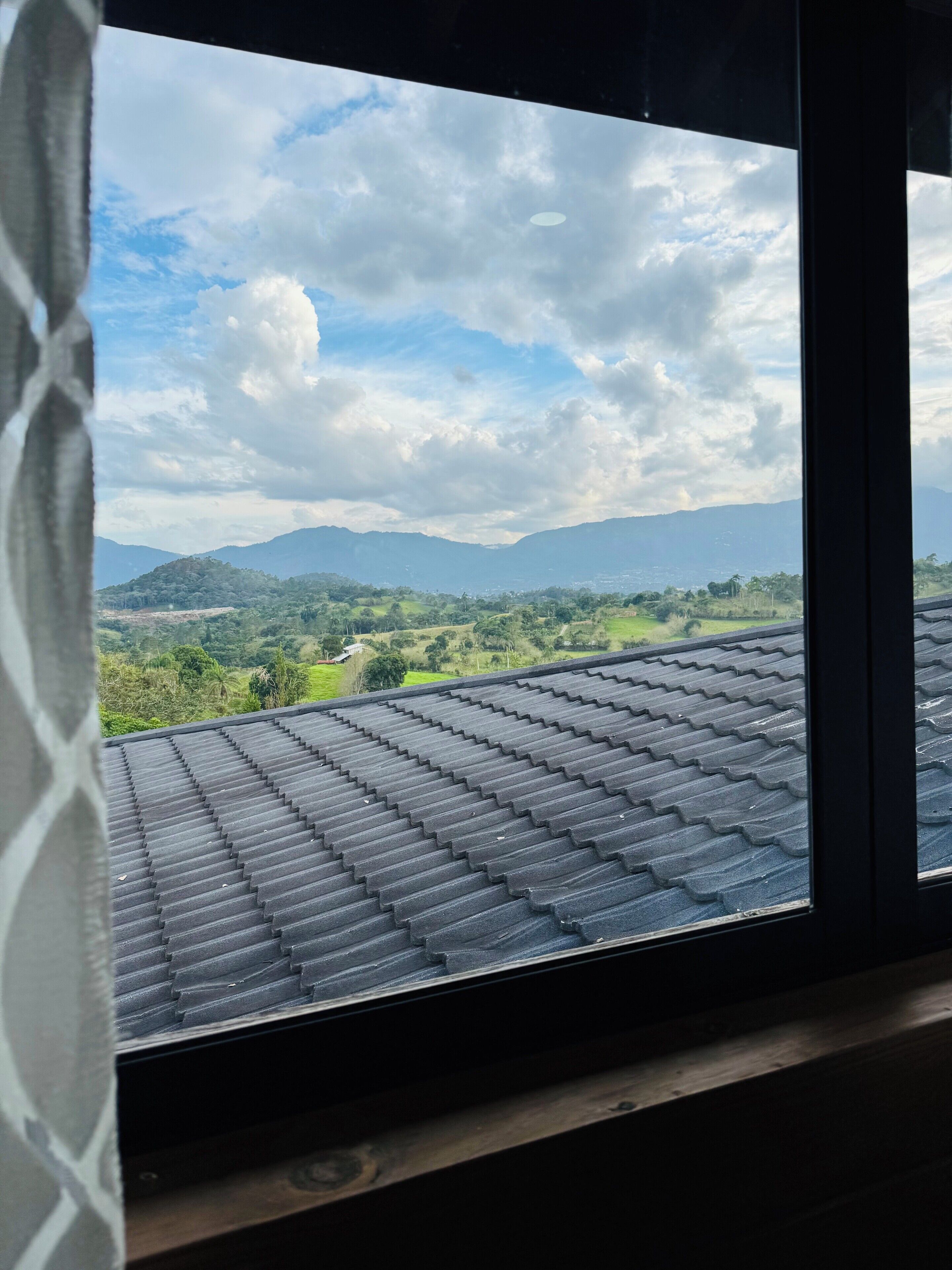 A view from a window with a tiled roof, overlooking a green landscape with hills and a partly cloudy sky.