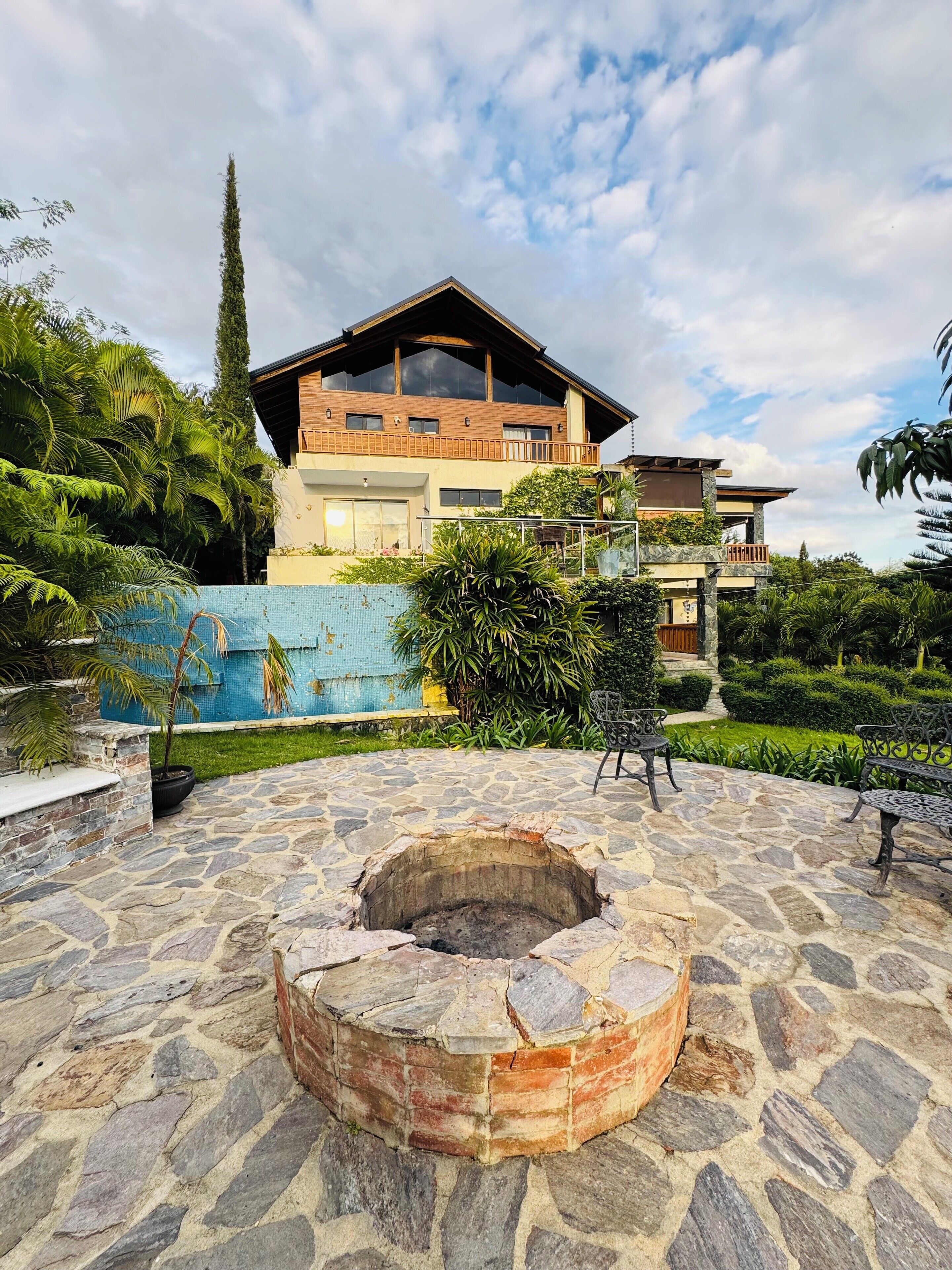 A stone patio with a fire pit, surrounded by lush greenery and a two-story building with a balcony.