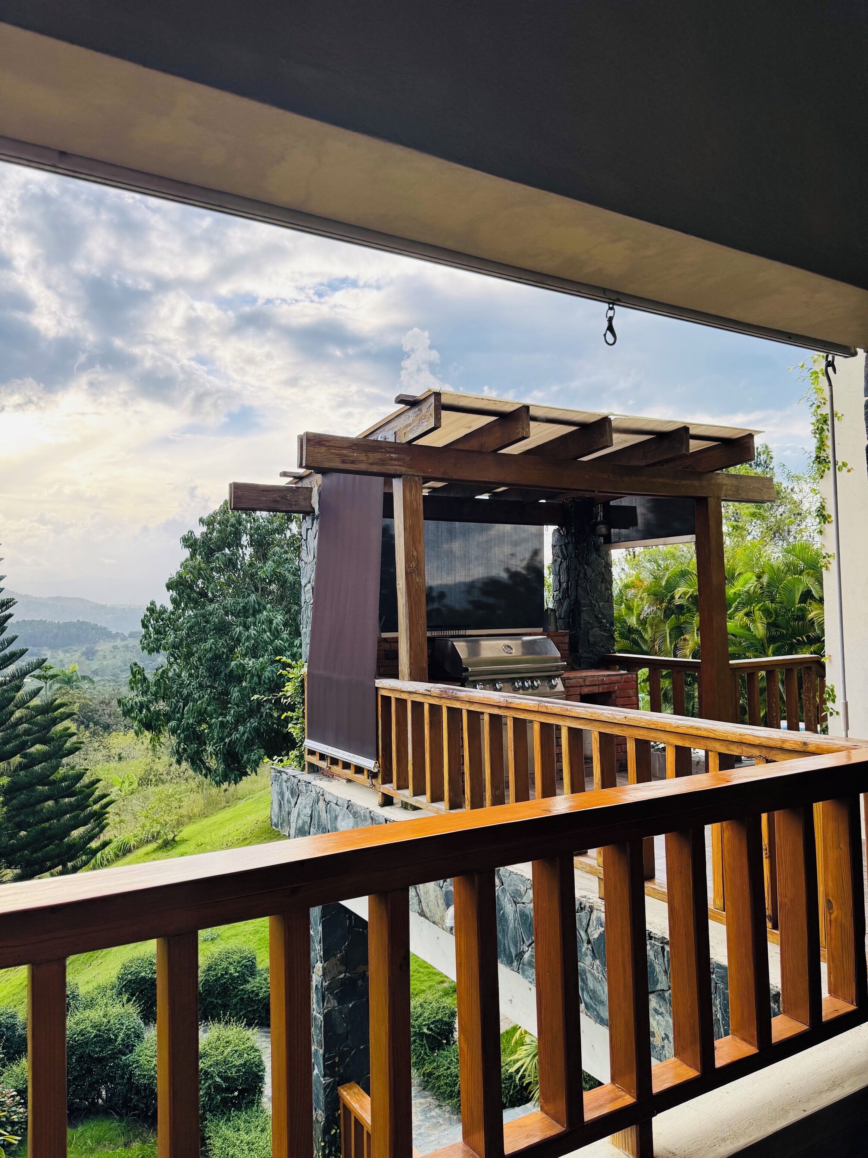Una terraza de madera con una parrilla y un televisor, con vista a un paisaje exuberante con árboles y una cadena montañosa distante.
