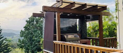 Una terraza de madera con una parrilla y un televisor, con vista a un paisaje exuberante con árboles y una cadena montañosa distante.