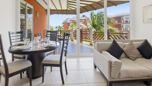 Ein Zimmer mit Esstisch für zwei Personen, einer Couch und einer Schiebetür zu einem Balkon mit Blick auf Gebäude und Palmen.