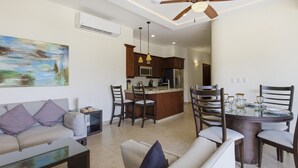 Ein Wohnzimmer mit einer Couch, einem Couchtisch und einem Essbereich mit Tisch und Stühlen. Im Hintergrund ist eine Küche mit Kühlschrank und Spüle zu sehen.