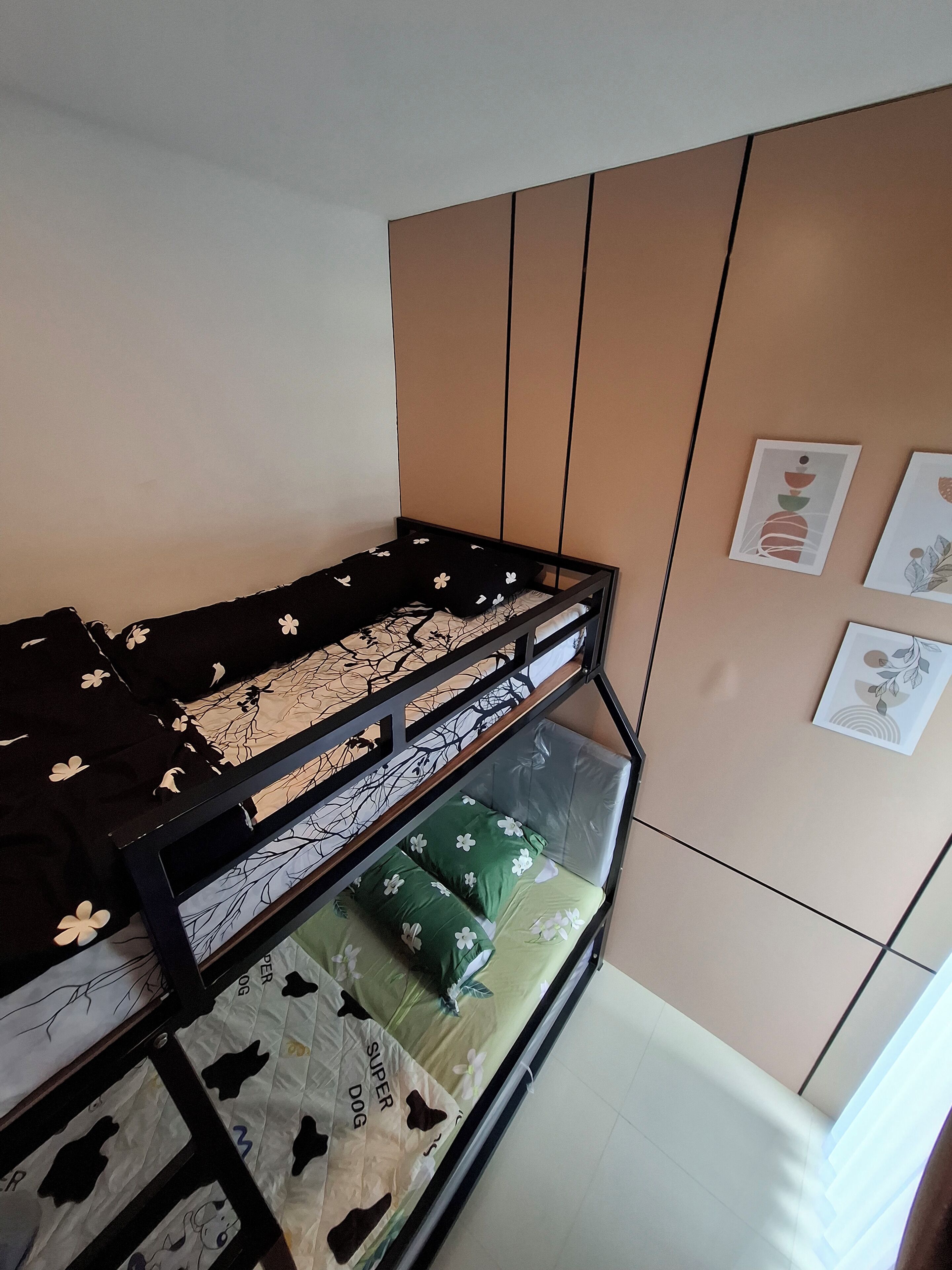 A bunk bed with a lower bunk covered in a floral-patterned sheet and a top bunk with a black sheet. There are wall-mounted pictures and a window with curtains.